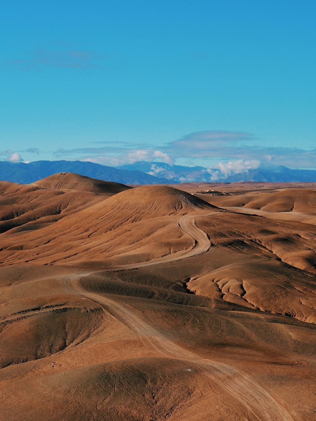 A dirt track winding between the bare, rounded brown hills of the Agafay desert, with the blue ridges of the Atlas Mountains on the horizon.