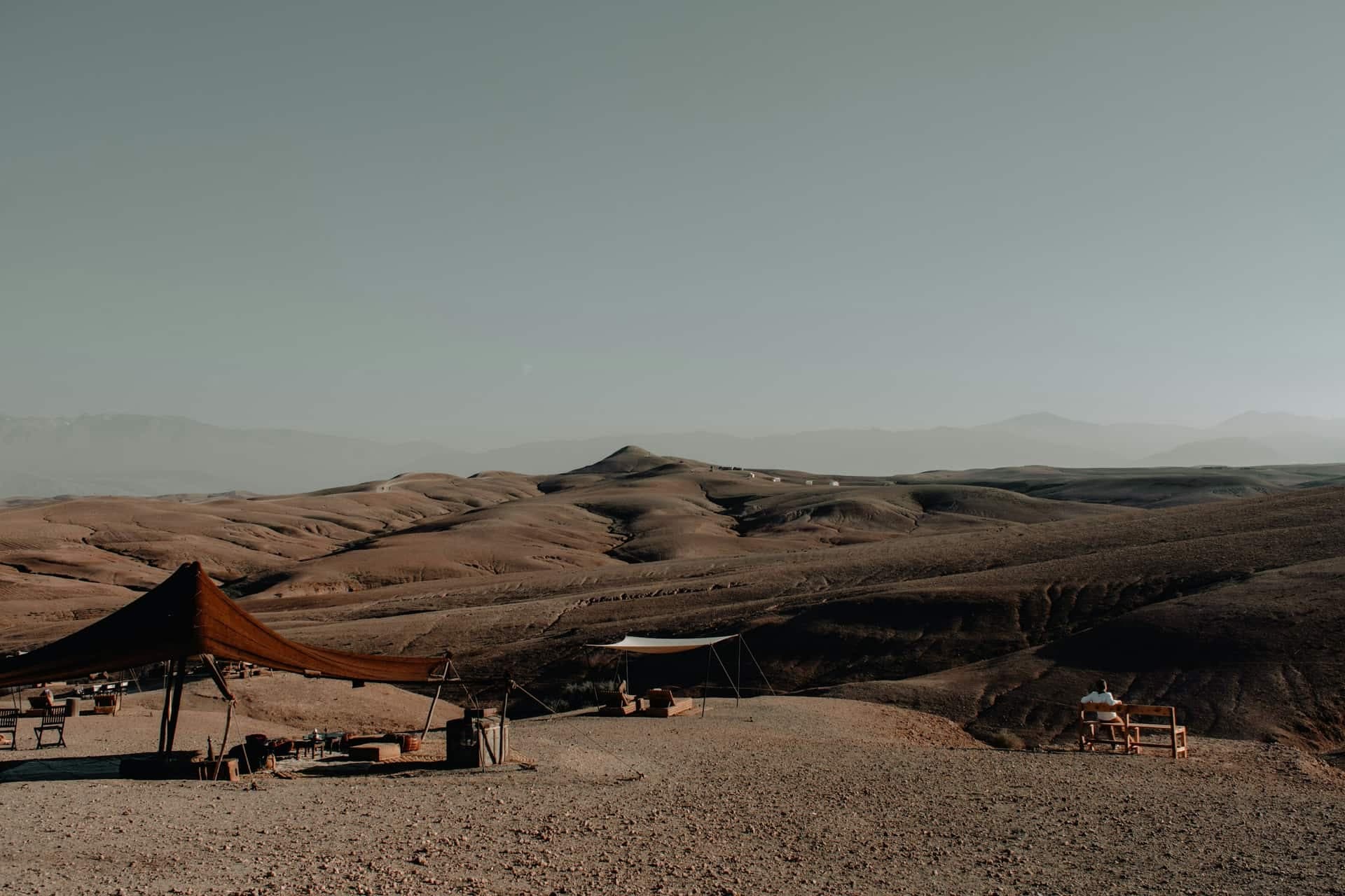 A shade canopy and low seating on stony ground at the edge of the Agafay desert, looking out over rolling brown hills towards distant mountains in haze.