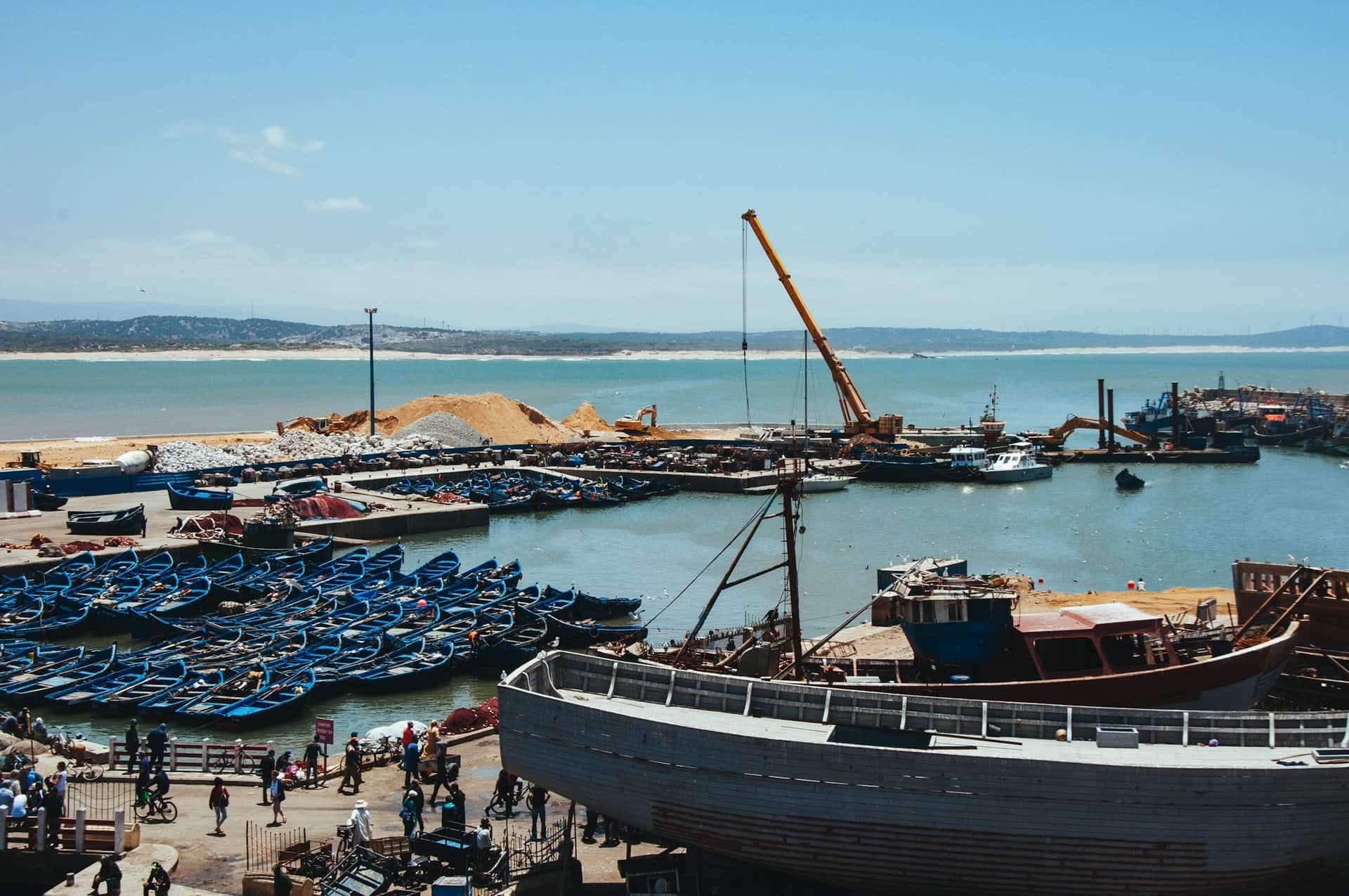 The fishing port of Essaouira, with rows of blue wooden boats moored inside the harbour wall, a boat under construction on the quay and the Atlantic beyond.
