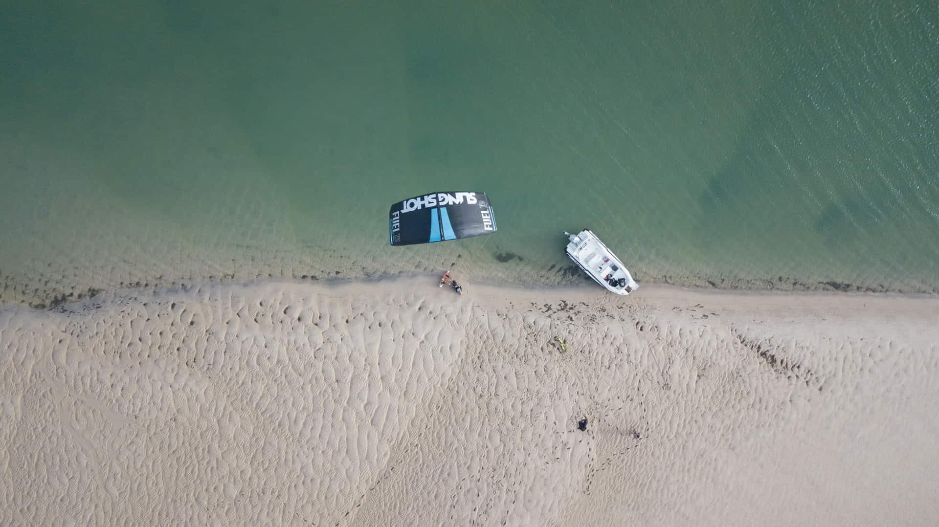 Aerial view of the shallow green water and sand of the Dakhla lagoon, with a kite and a small white boat on the shore.