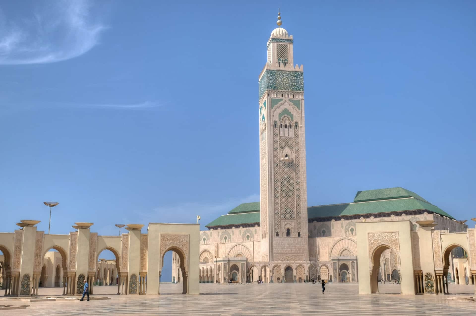 The minaret of the Hassan II Mosque in Casablanca rising above the arcaded esplanade and green-tiled roofs, under a clear sky.