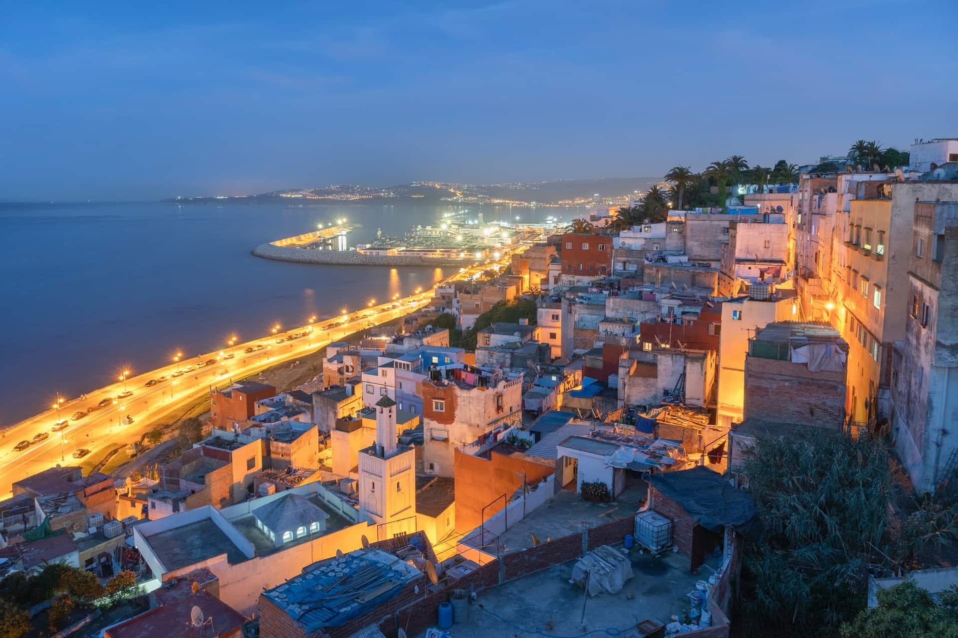 Tangier at dusk from above, the lit medina and seafront road curving along the bay towards the port.