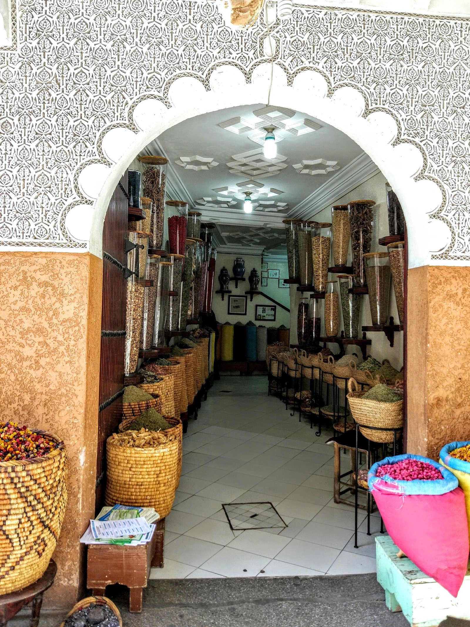 A spice and herbalist shop in the Marrakech medina seen through a carved white arch, with jars on wooden shelves and baskets of dried roses at the door.