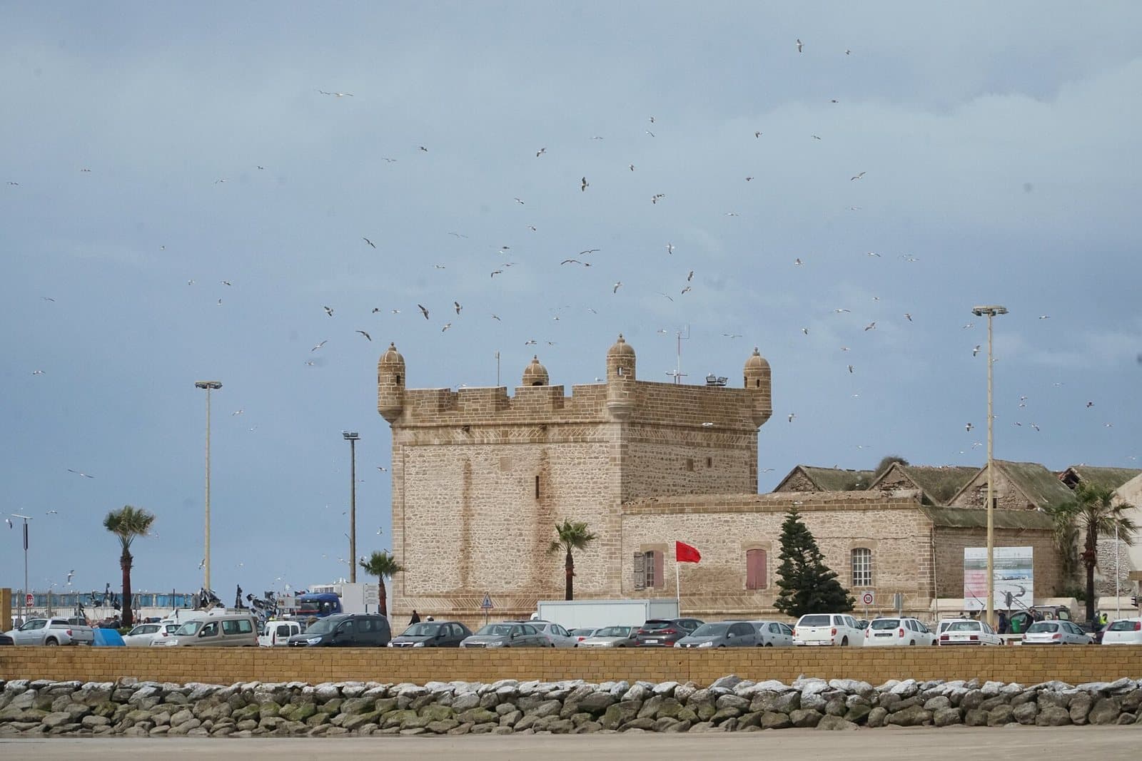 The square stone bastion of the Skala du Port in Essaouira under a grey sky, with gulls wheeling above and a breakwater in front.