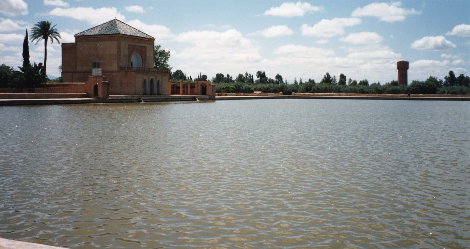 The green-roofed Menara pavilion reflected at the far end of its wide water basin, with palms and olive trees under a blue sky.