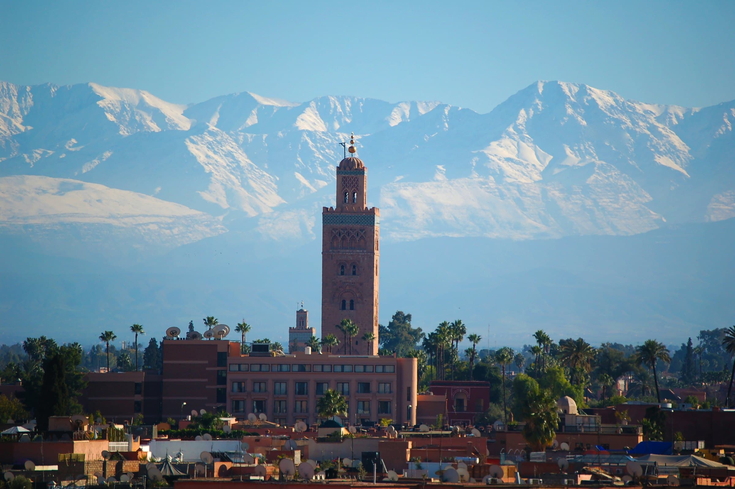 The Koutoubia minaret rising above the rooftops of Marrakech, with the snow-covered peaks of the High Atlas behind the city.