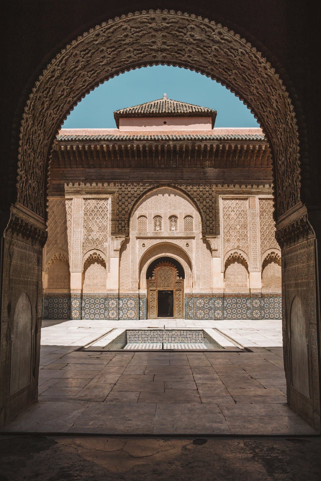 The courtyard of the Ben Youssef Medersa in Marrakech, seen through a carved archway, with zellige tilework and a still central pool.