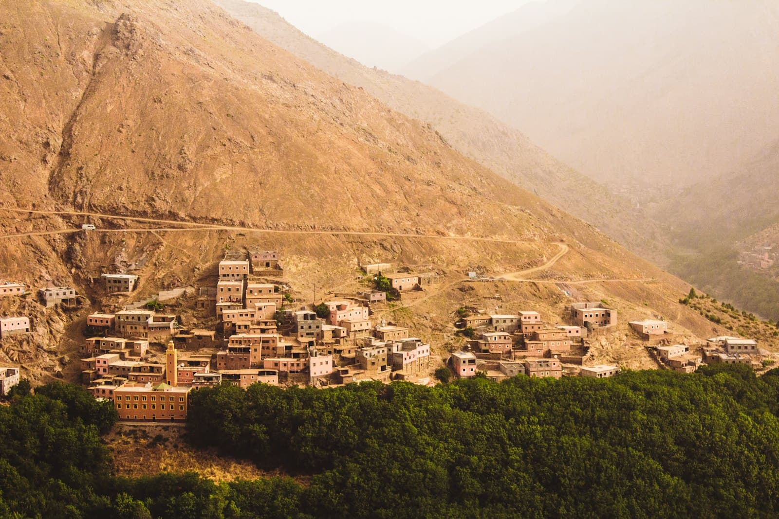 A High Atlas village of earth-built houses on a mountainside above a wooded valley, with the access road visible cut into the slope.