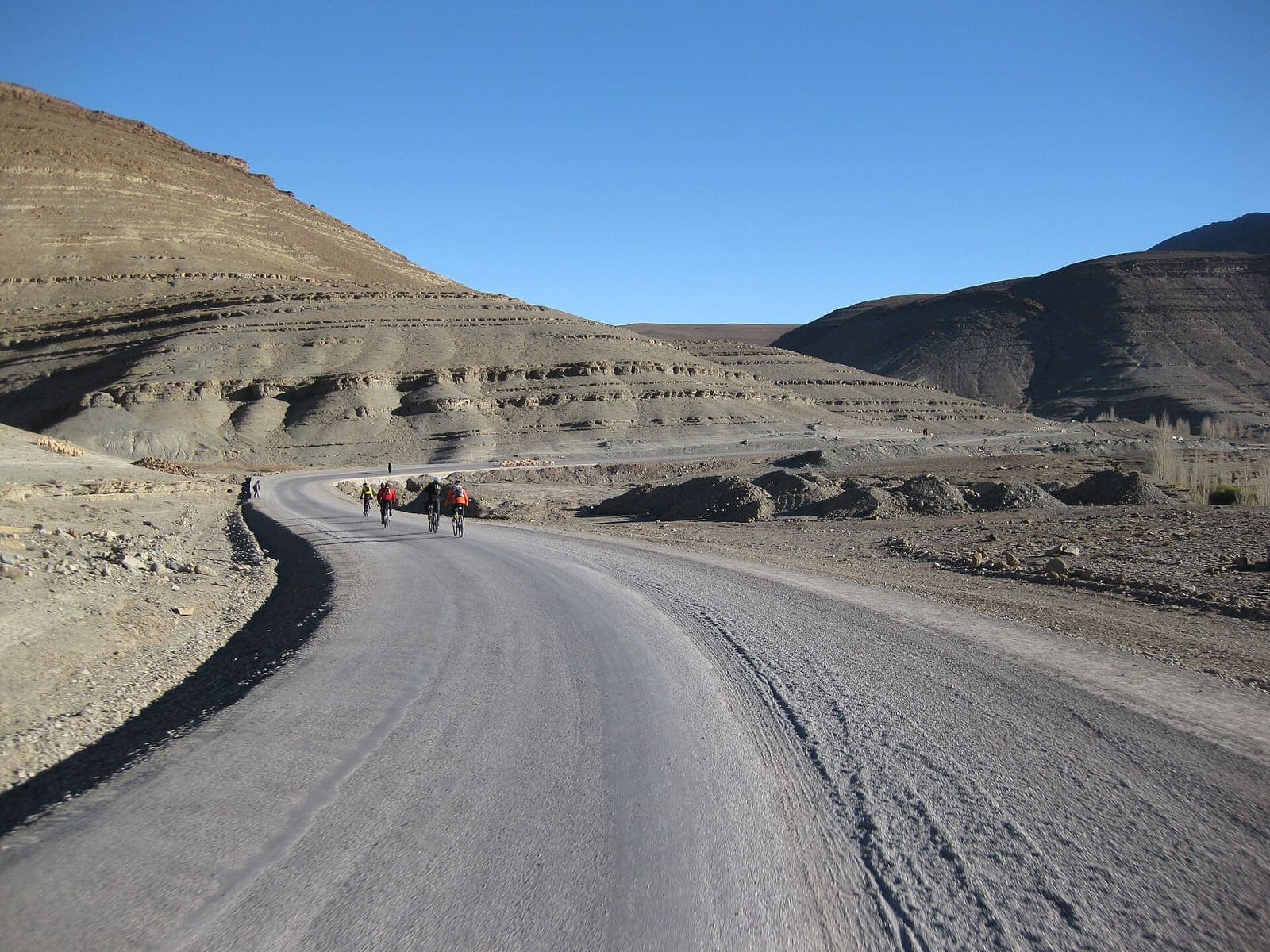 A small group of cyclists far along a winding mountain road between bare, layered hills in the Atlas, under a clear blue sky.