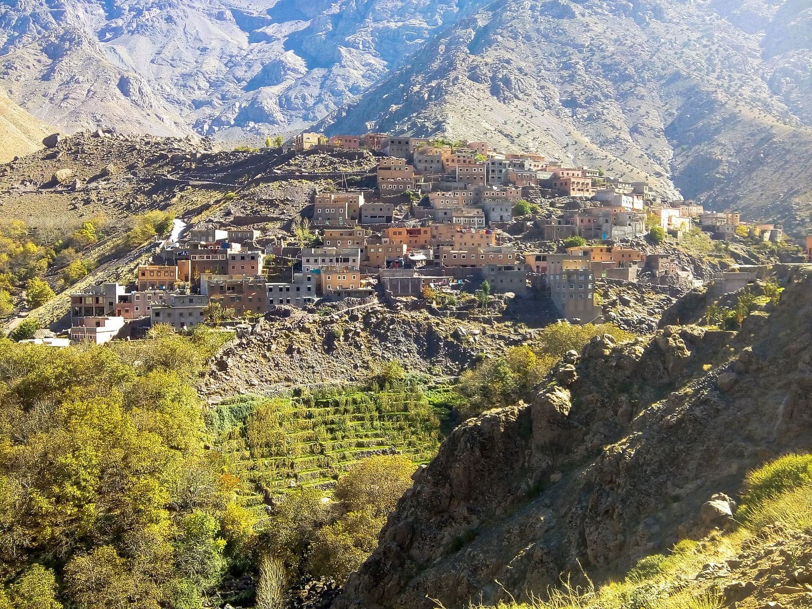An Amazigh village of stacked earth and stone houses on a High Atlas slope above terraced fields and autumn trees near Imlil.