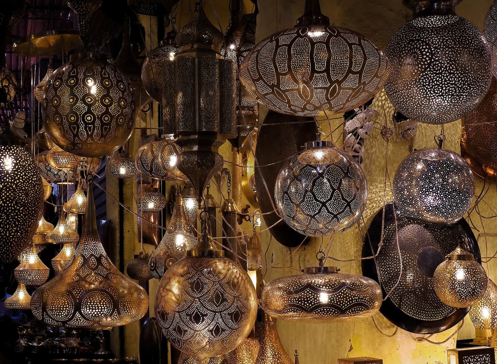 Pierced brass and copper lanterns hanging close together in a stall in Souk Haddadine, glowing against the dark.