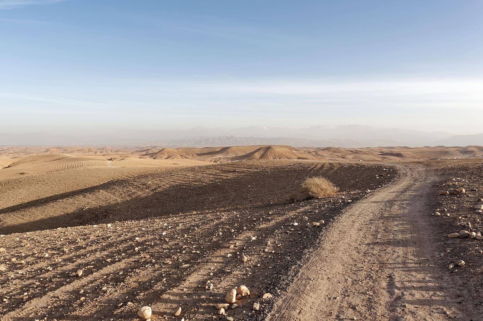 A rough dirt track running across the stony Agafay desert towards low hills, with the High Atlas faint on the horizon.