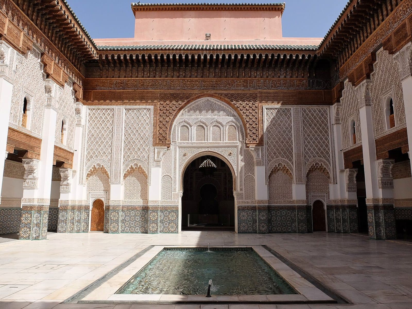 The main courtyard of the Ben Youssef Madrasa, with carved stucco and cedar walls, zellige tiling and a long marble pool before the prayer hall arch.