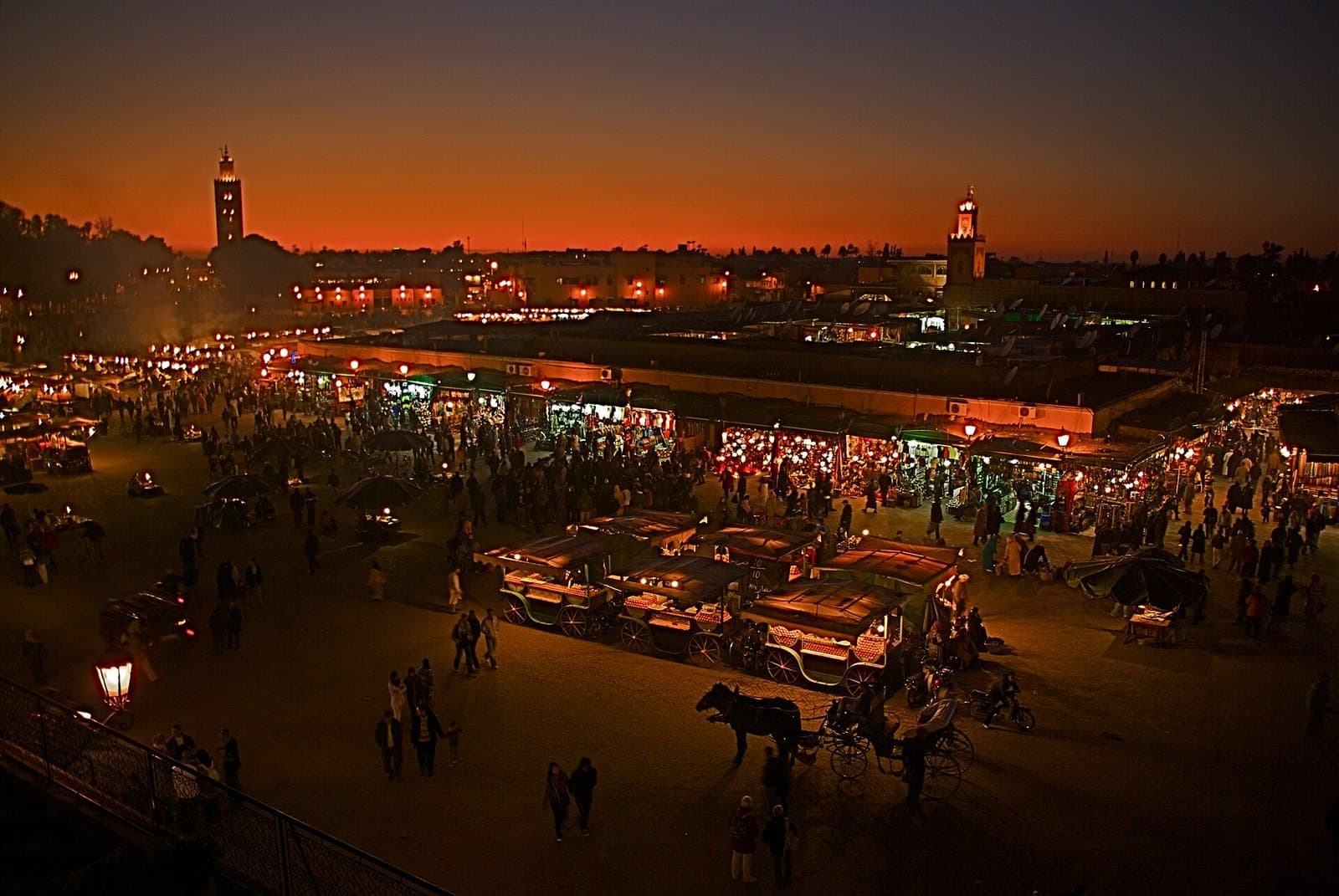 Jemaa el-Fna square seen from above at dusk, its lit stalls and juice carts glowing under an orange sky with the Koutoubia minaret on the skyline.