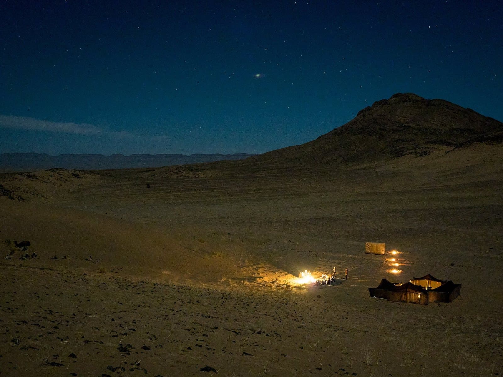 A small desert camp glowing with lamps at night on a wide stony plain below a dark hill, under a starry sky.