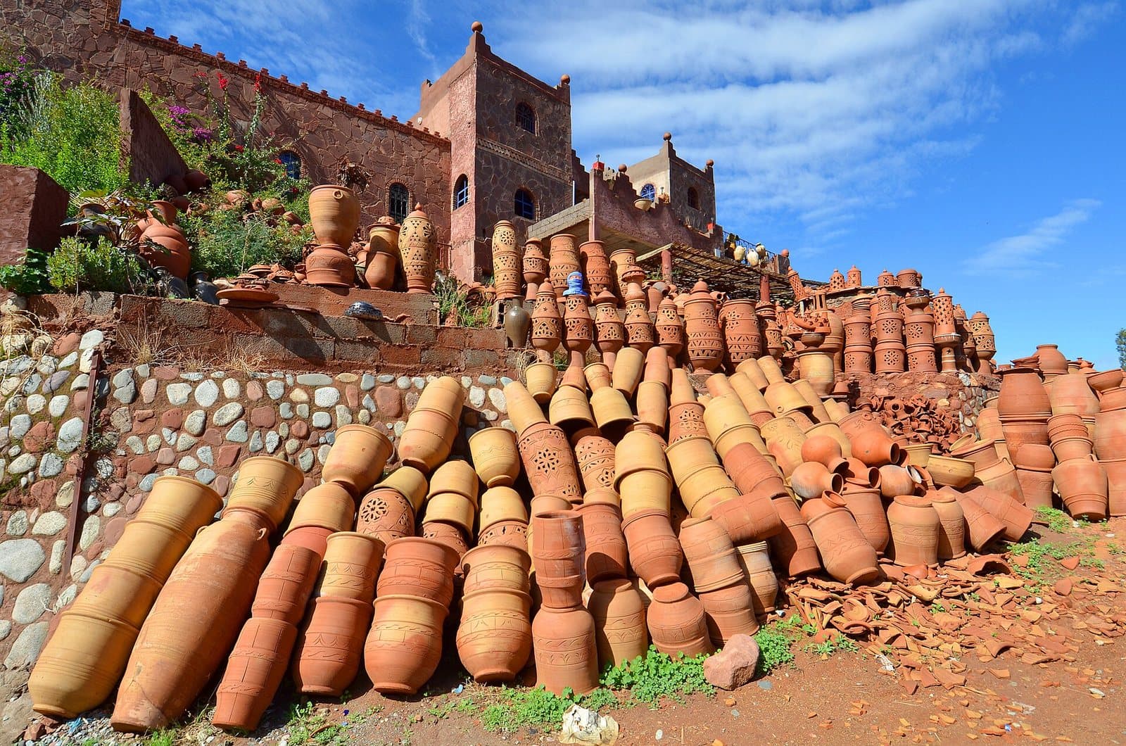 Hundreds of terracotta pots stacked on a stone terrace along the Ourika valley road, below an earthen kasbah tower.