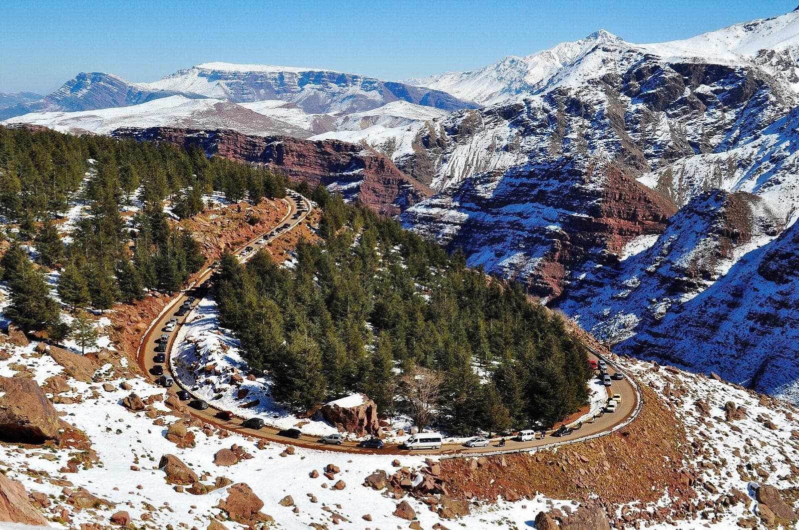 A mountain road winding around a pine-covered spur towards Oukaïmeden, below snow-covered High Atlas ridges.