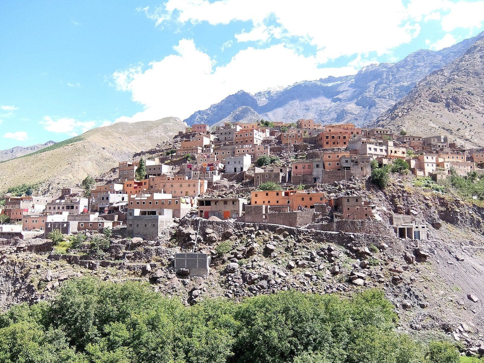 The stone and earth houses of Aroumd stacked on a rocky spur above green trees, with High Atlas ridges behind.